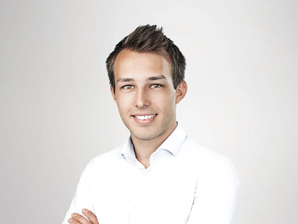 Ein junger Mann steht mit verschränkten Armen vor einem neutralen, hellen Hintergrund. Er trägt ein weißes Hemd und beigefarbene Hose und lächelt freundlich in die Kamera.