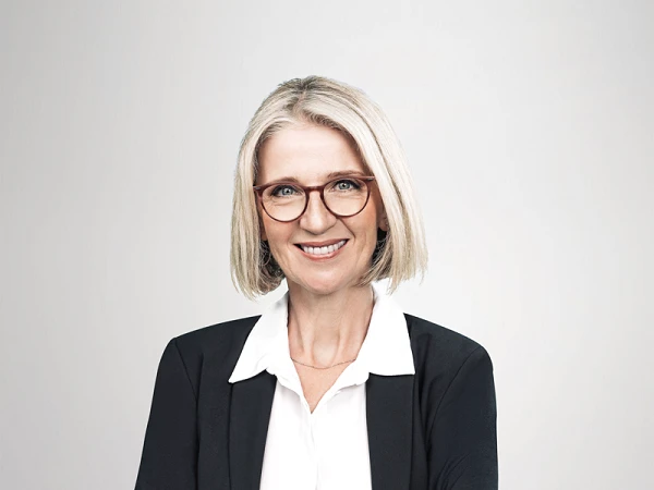 Eine selbstbewusste Frau mit blonden, schulterlangen Haaren, die eine elegante Brille trägt, steht mit verschränkten Armen in einem schwarzen Blazer und einer weißen Bluse vor einem hellen Hintergrund.
