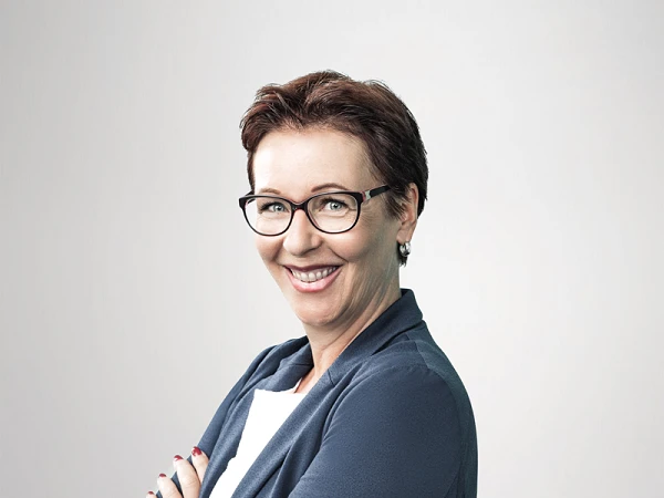 Eine lachende Frau mit kurzen, braunen Haaren und Brille steht mit verschränkten Armen. Sie trägt ein dunkelblaues Blazer und ein weißes Oberteil, vor einem hellen, einfarbigen Hintergrund.
