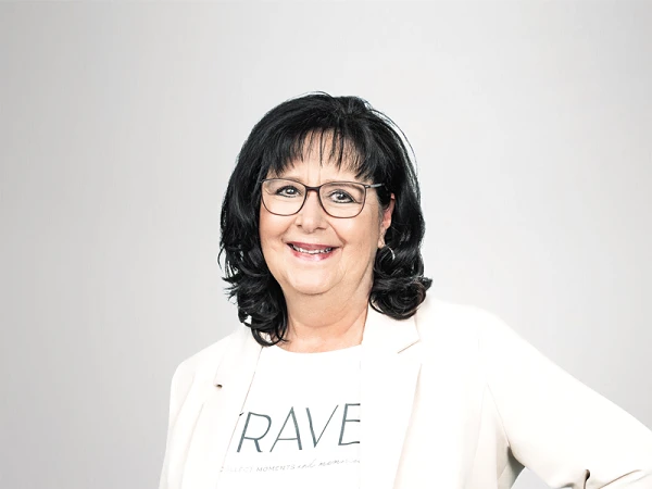 Die Frau steht lächelnd vor einem hellen, einfarbigen Hintergrund. Sie trägt ein helles Blazer-Jackett über einem T-Shirt mit dem Aufdruck "BRAVE" und eine auffällige Hose mit Pfauenfedern-Muster. Sie hat lange, dunkle Haare und eine Brille.