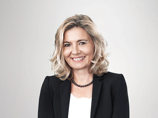 Eine lächelnde Frau mit schulterlangen blonden Haaren trägt einen schwarzen Blazer über einem weißen Oberteil und hat die Arme vor der Brust verschränkt. Der Hintergrund ist hell und neutral.