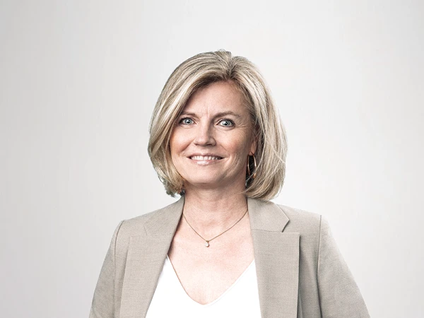 Eine lächelnde Frau mit blonden, schulterlangen Haaren trägt einen grauen Blazer über einem weißen Oberteil und Jeans. Sie steht mit den Händen in den Taschen und hat einen selbstbewussten Ausdruck. Der Hintergrund ist hell und neutral.
