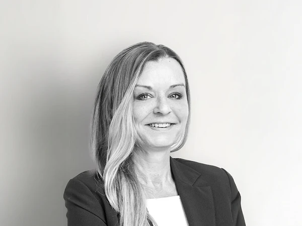 Eine Frau mit langen, glatten Haaren und einem freundlichen Lächeln trägt einen Blazer und steht vor einem hellen, einfarbigen Hintergrund.