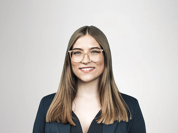 Eine Frau mit langen, glatten Haaren und Brille steht lächelnd vor einem hellen, neutralen Hintergrund. Sie trägt einen dunkelblauen Blazer über einem schwarzen Oberteil und hat eine Uhr an ihrem Handgelenk.