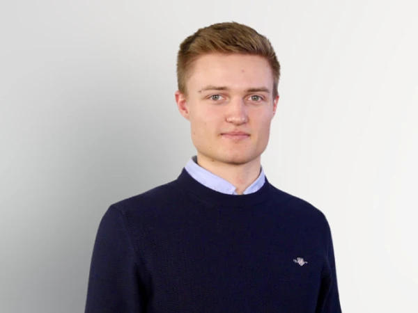 Ein junger Mann trägt einen dunkelblauen Pullover über einem hellblauen Hemd und steht mit verschränkten Händen vor einem hellen, einfarbigen Hintergrund.