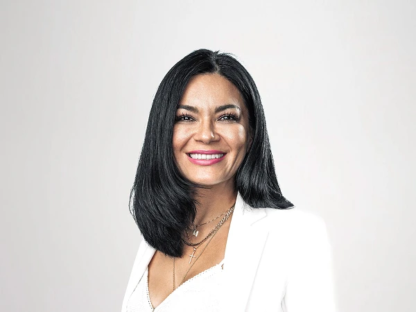 Eine Frau mit langen schwarzen Haaren trägt ein elegantes, weißes Kleidungsset bestehend aus einer Bluse und einem Blazer. Sie lächelt freundlich und posiert mit beiden Händen in den Hosentaschen, vor einem hellen, neutralen Hintergrund.
