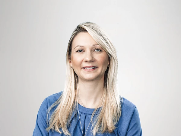 Eine lächelnde Frau mit langen, blonden Haaren trägt ein blaues Kleid und steht mit verschränkten Armen vor einem neutralen, hellen Hintergrund.