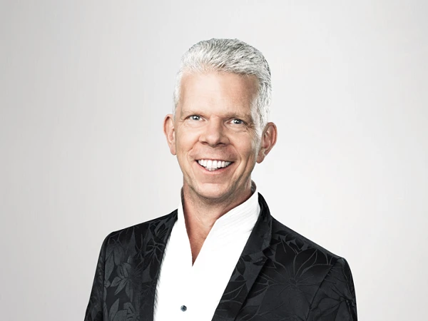 Ein lächelnder Mann in einem eleganten, schwarz gemusterten Anzug mit einem weißen Hemd. Der Hintergrund ist hell und neutral gehalten.