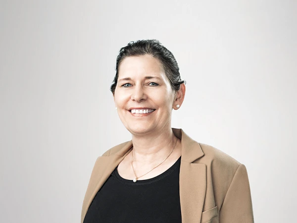 Eine lächelnde Frau in einem zweitönigen Outfit, bestehend aus einem schwarzen Top und einem beigen Blazer, steht vor einem neutralen Hintergrund und posiert mit verschränkten Fingern auf Höhe ihrer Taille.