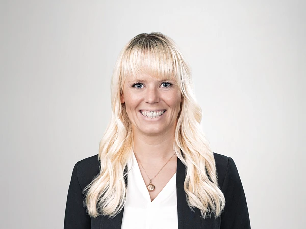 Eine lächelnde Frau mit langen blonden Haaren trägt ein schwarzes Blazerjackett und ein weißes Oberteil. Sie steht mit geschlossenen Händen vor ihrem Körper und hat einen neutralen hellen Hintergrund.