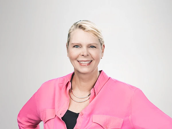 Eine blonde Frau lächelt selbstbewusst in einem leuchtend pinken Hemd und schwarzer Hose, ihre Hände auf den Hüften positioniert.