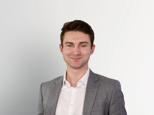 Ein junger Mann in einem grauen Blazer und heller Hemd steht mit verschränkten Armen vor einem hellen, einfarbigen Hintergrund und lächelt.