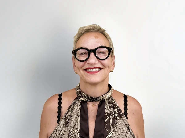 Eine lächelnde Frau mit kurzen, blonden Haaren trägt eine elegante Bluse mit einem auffälligen Muster und große, schwarze Brillen. Der Hintergrund ist hell und neutral.