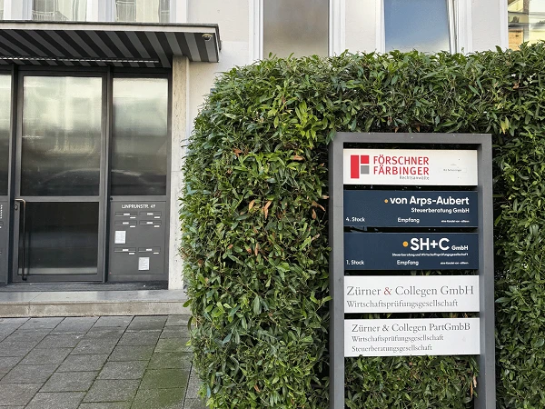 Das Bild zeigt ein modernes Gebäude mit einer Glasfront und einem Empfangsbereich sowie einem Pflanzenbeet im Vordergrund. An einem Schilderständer sind mehrere Firmennamen und ihre Kontaktdaten angezeigt, darunter "Förschner Farbinger", "von Arps-Aubert", "SH+C" und "Zirner & Collegen GmbH".