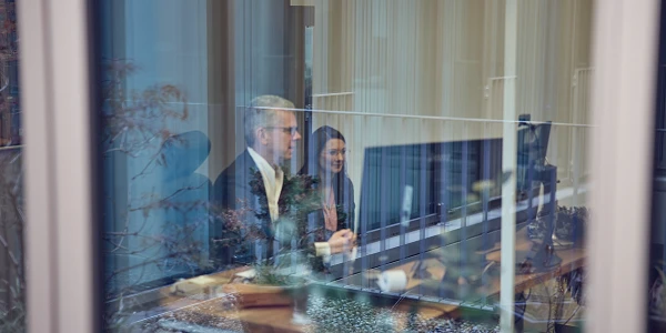 Zwei Personen sitzen an einem Tisch vor einem Computer in einem modernen Büro, während sie durch ein Fenster beobachtet werden. Im Vordergrund sind Pflanzen und der Büroausblick sichtbar.