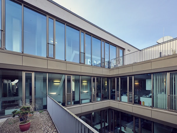 Moderne Architektur mit großen Fensterflächen und einem Innenhof, der von mehreren Etagen umgeben ist. Im Vordergrund sind kleine Pflanzen und ein Kiesbereich zu sehen.