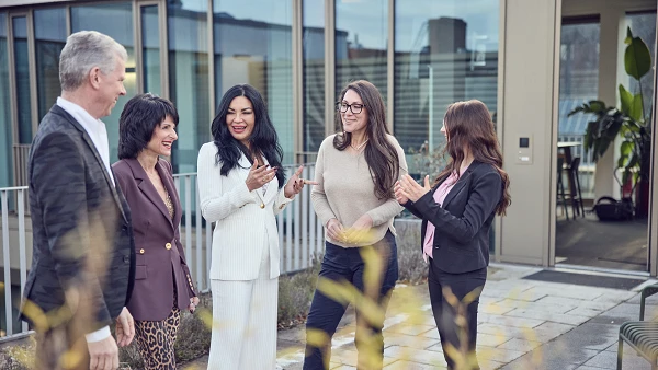 Fünf Personen stehen in einer modernen, lichtdurchfluteten Umgebung und interagieren miteinander, während sie lächeln und applaudieren. Die Frauen tragen verschiedene stilvolle Outfits, während die Männer formelle Kleidung wählen. Im Hintergrund sind große Fenster und Pflanzen sichtbar.