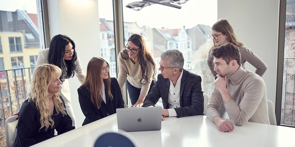 Eine Gruppe von sieben Personen versammelt sich um einen Tisch mit einem Laptop in einem modernen Büroraum, während sie zusammen an einem Projekt arbeiten und sich gegenseitig austauschen. Im Hintergrund sind Fenster mit Blick auf eine städtische Umgebung zu sehen.