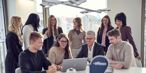 Eine Gruppe von acht Personen in einem modernen Büroraum, die um einen Tisch versammelt sind und zusammen an einem Laptop arbeiten, während einige lächeln und andere aufmerksam schauen. Im Hintergrund sind Fenster mit Stadtblick zu sehen.