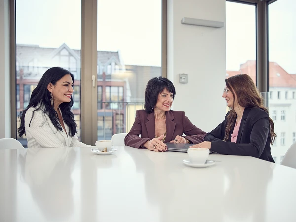 Drei Frauen sitzen an einem hellen Tisch in einem modernen Büroraum, während sie sich unterhalten. Vor ihnen stehen Kaffeetassen, und eine von ihnen hat einen Laptop geöffnet. Durch die großen Fenster ist der Blick auf städtische Architektur zu sehen.