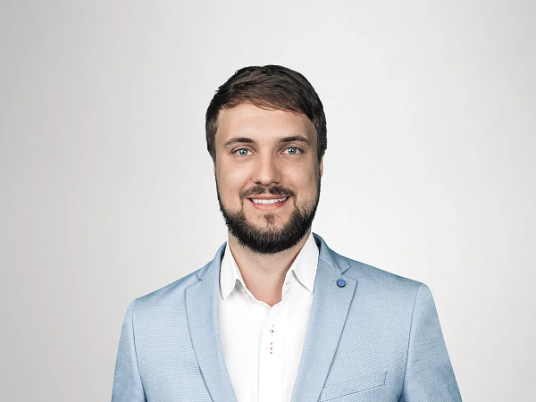 Ein Mann mit einem freundlichen Lächeln trägt einen hellblauen Anzug und steht mit beiden Händen in den Taschen vor einem neutralen, hellen Hintergrund.