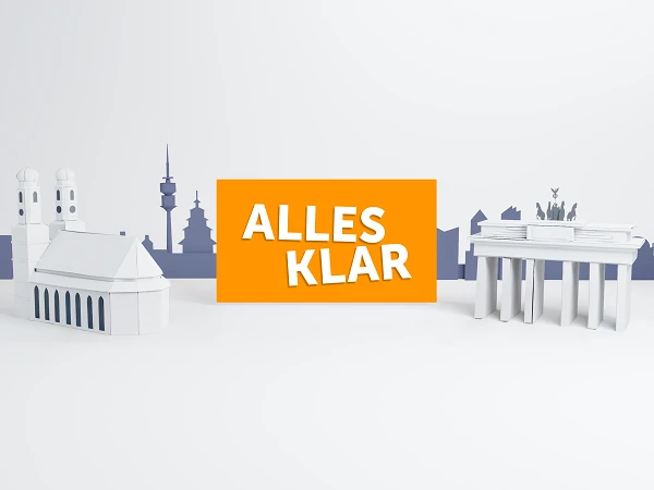 Zwei stilisierte, weiße Architekturmodelle, darunter ein bekanntes Wahrzeichen, das als Brandenburger Tor identifiziert werden kann, stehen auf einem hellen Hintergrund. Zwischen den Gebäuden liegt ein leuchtend orangefarbener Schriftzug mit dem Text "ALLES KLAR". Im Hintergrund sind schwache Silhouetten weiterer Gebäude zu sehen.
