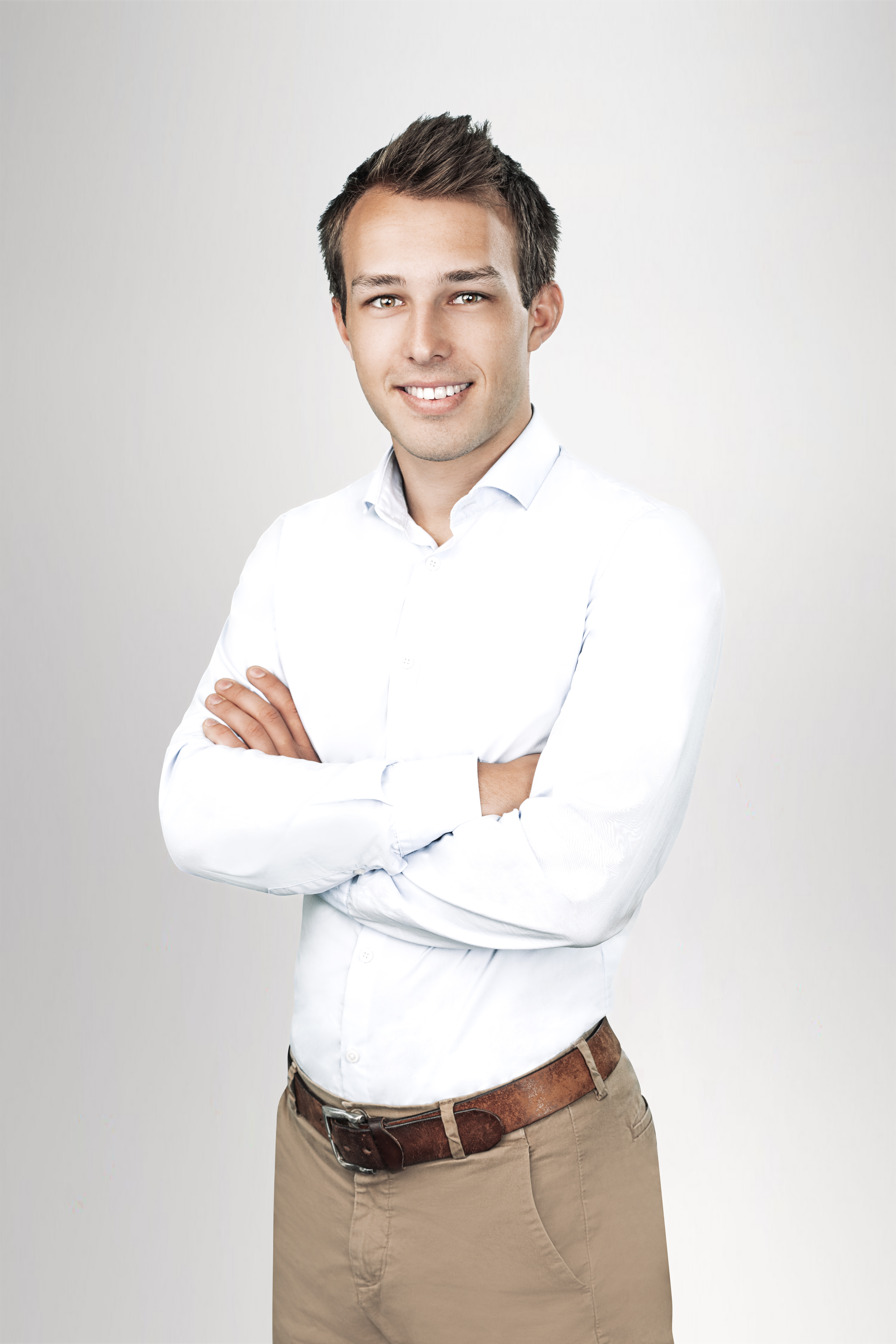 Ein junger Mann steht mit verschränkten Armen vor einem neutralen, hellen Hintergrund. Er trägt ein weißes Hemd und beigefarbene Hose und lächelt freundlich in die Kamera.
