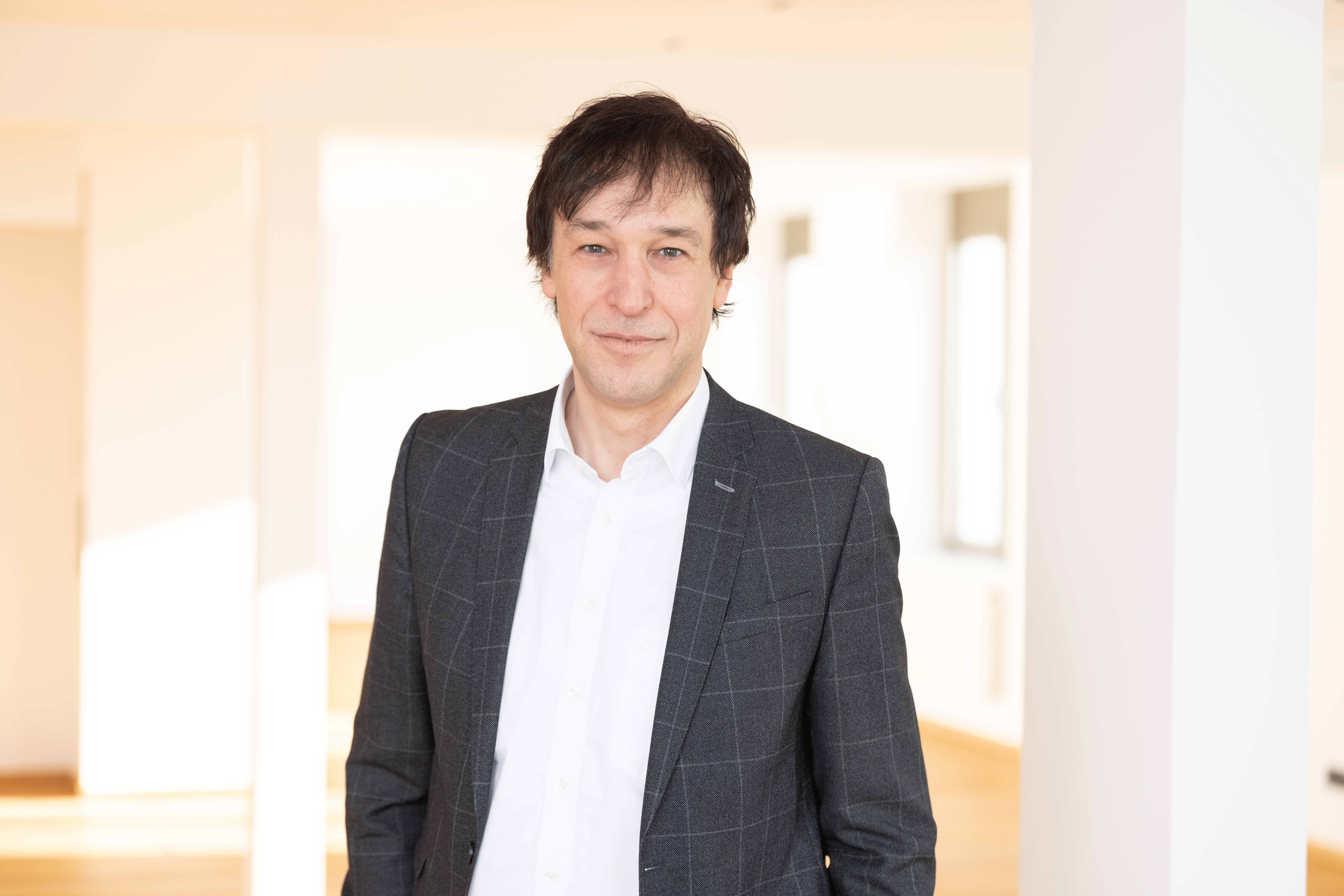 Ein Mann mit dunklen, leicht gewellten Haaren trägt einen grauen Anzug mit karierter Textur und ein weißes Hemd. Er steht in einem hellen Raum mit großen Fenstern und Holzboden, lächelt leicht und schaut in die Kamera.