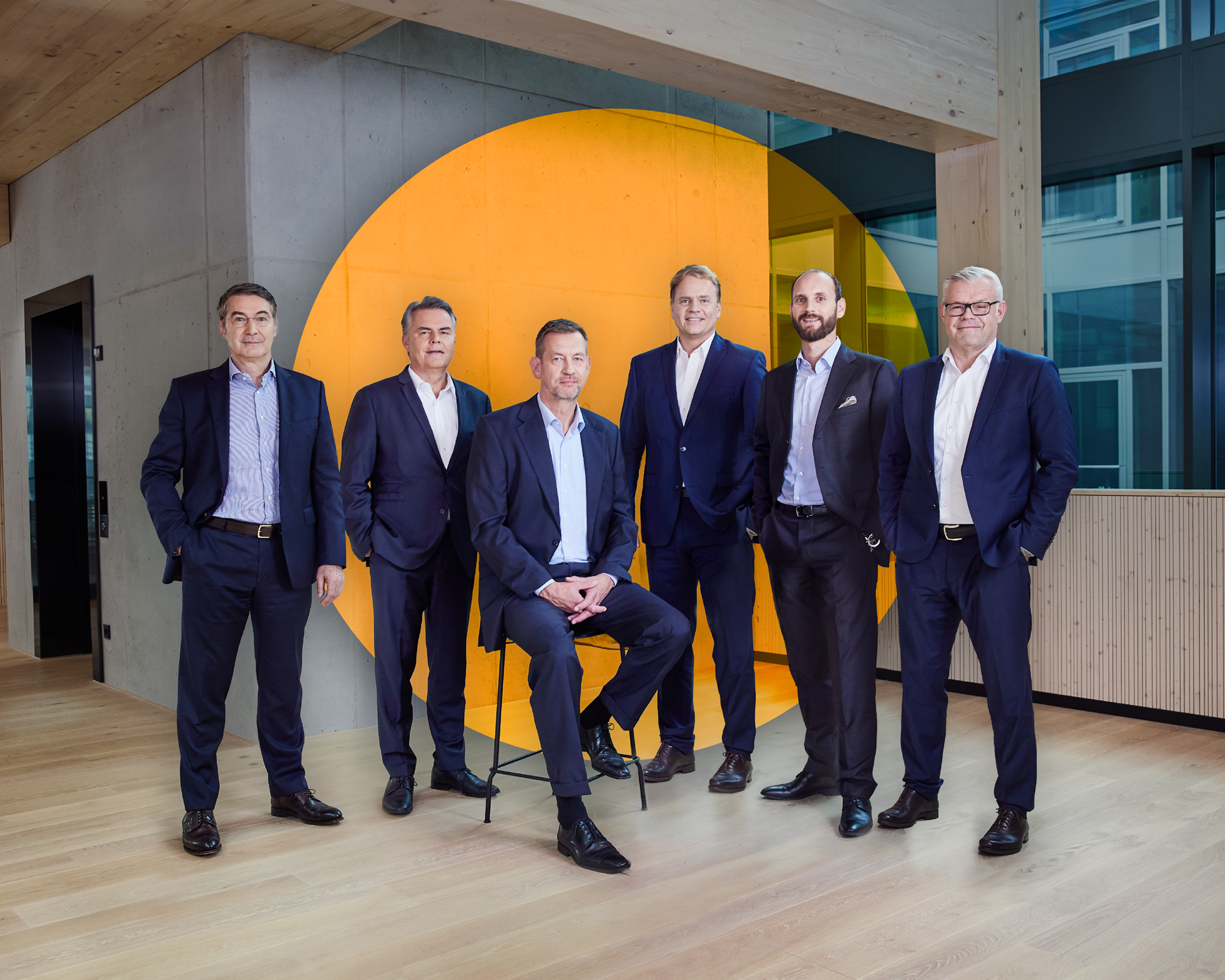 Sechs Männer in Anzügen stehen und sitzen in einem modernen Büroumfeld vor einer großen, orangefarbenen Wandinstallation. Sie posieren in einer Reihe, während sie in die Kamera schauen.