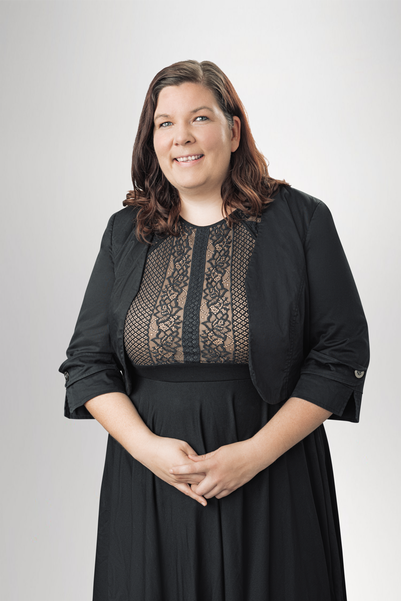 Eine Frau mit lockigem, braunem Haar lächelt freundlich in die Kamera, bekleidet mit einem schwarzen Oberteil mit Spitzenakzenten und einem schwarzen Rock. Sie steht mit verschränkten Händen vor einem neutralen Hintergrund.