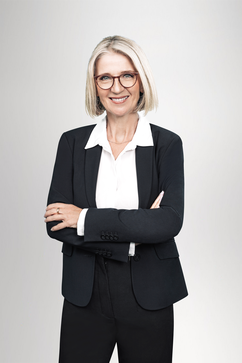 Eine selbstbewusste Frau mit blonden, schulterlangen Haaren, die eine elegante Brille trägt, steht mit verschränkten Armen in einem schwarzen Blazer und einer weißen Bluse vor einem hellen Hintergrund.
