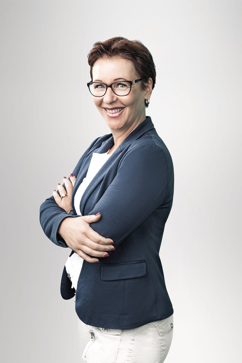 Eine lachende Frau mit kurzen, braunen Haaren und Brille steht mit verschränkten Armen. Sie trägt ein dunkelblaues Blazer und ein weißes Oberteil, vor einem hellen, einfarbigen Hintergrund.