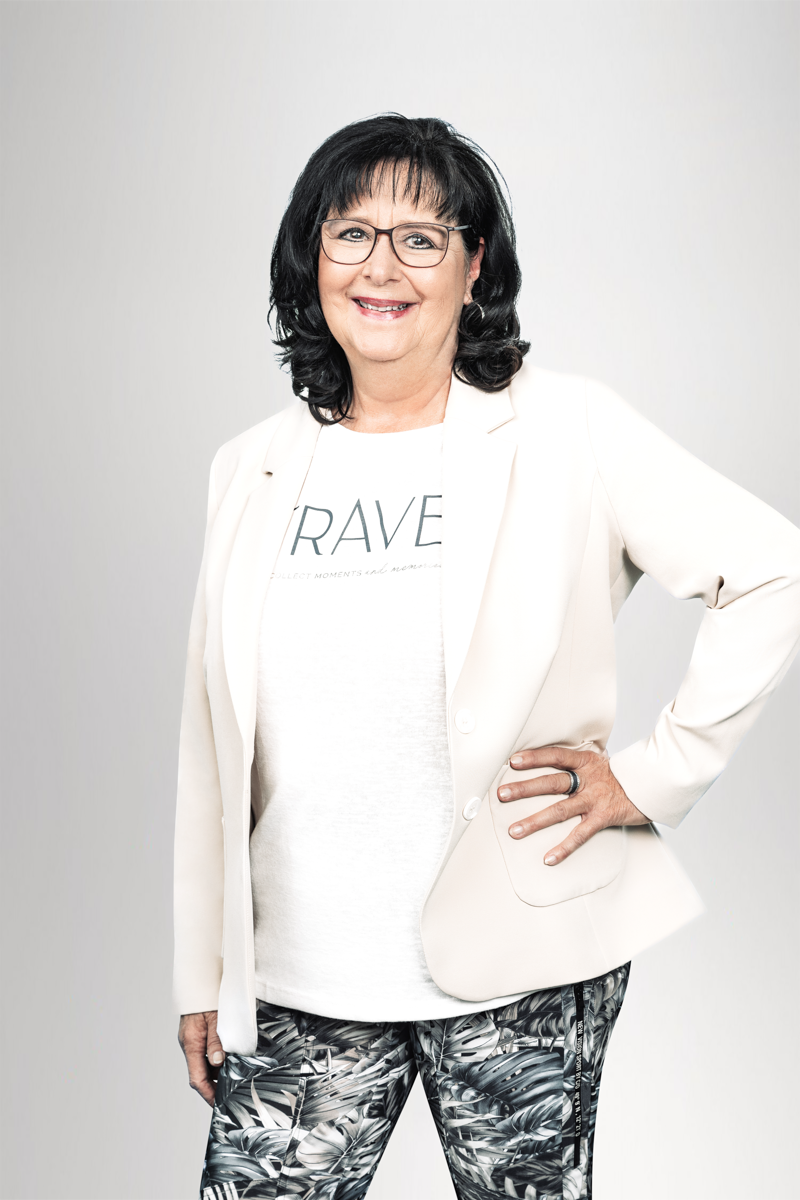 Die Frau steht lächelnd vor einem hellen, einfarbigen Hintergrund. Sie trägt ein helles Blazer-Jackett über einem T-Shirt mit dem Aufdruck "BRAVE" und eine auffällige Hose mit Pfauenfedern-Muster. Sie hat lange, dunkle Haare und eine Brille.