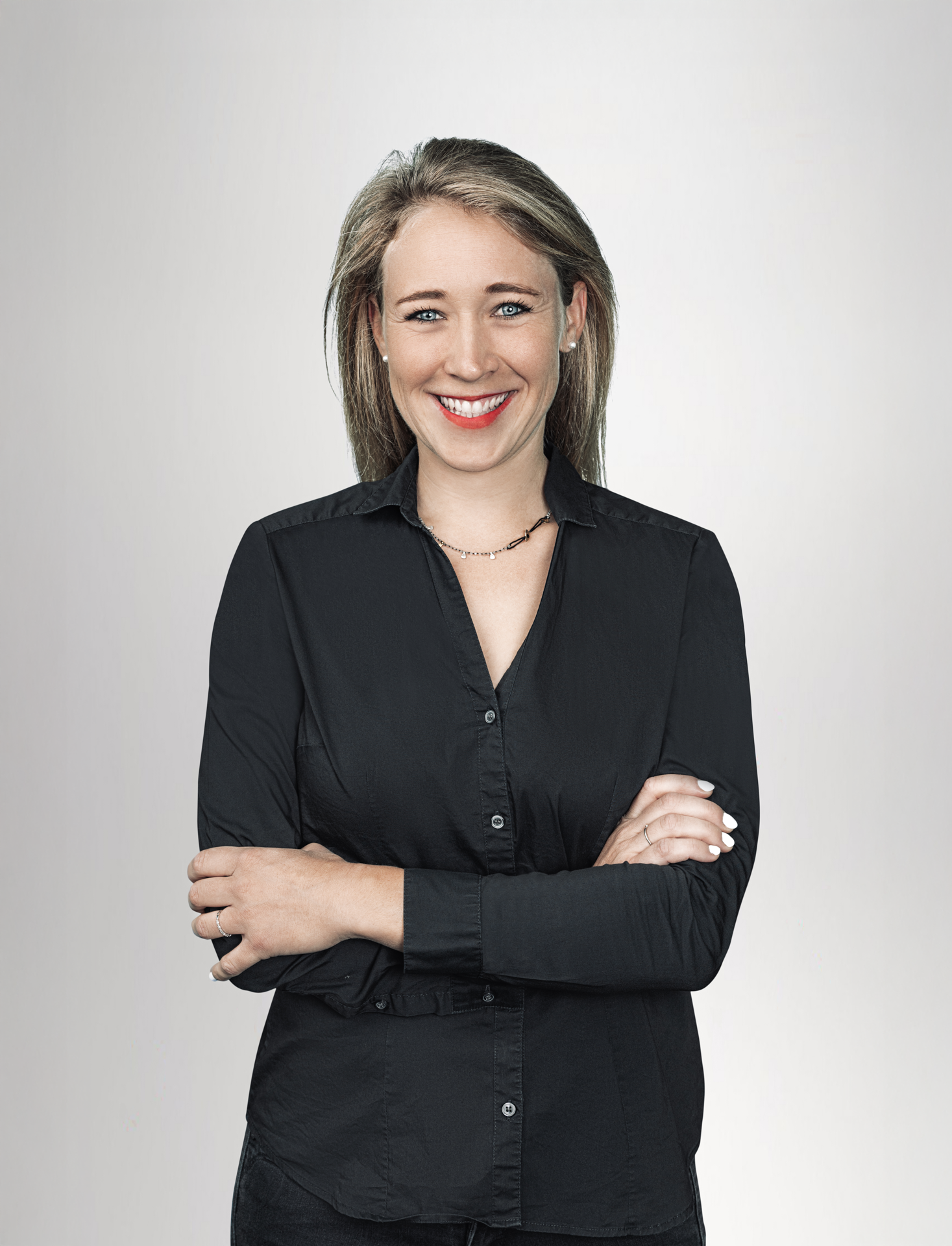 Eine lächelnde Frau mit langen, blonden Haaren steht mit verschränkten Armen vor einem neutralen, hellen Hintergrund. Sie trägt ein schwarzes Hemd und hat einen freundlichen Ausdruck.