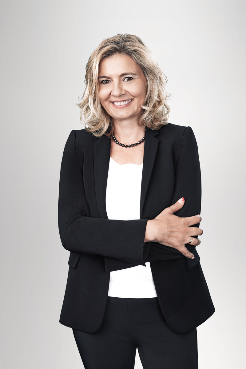 Eine lächelnde Frau mit schulterlangen blonden Haaren trägt einen schwarzen Blazer über einem weißen Oberteil und hat die Arme vor der Brust verschränkt. Der Hintergrund ist hell und neutral.