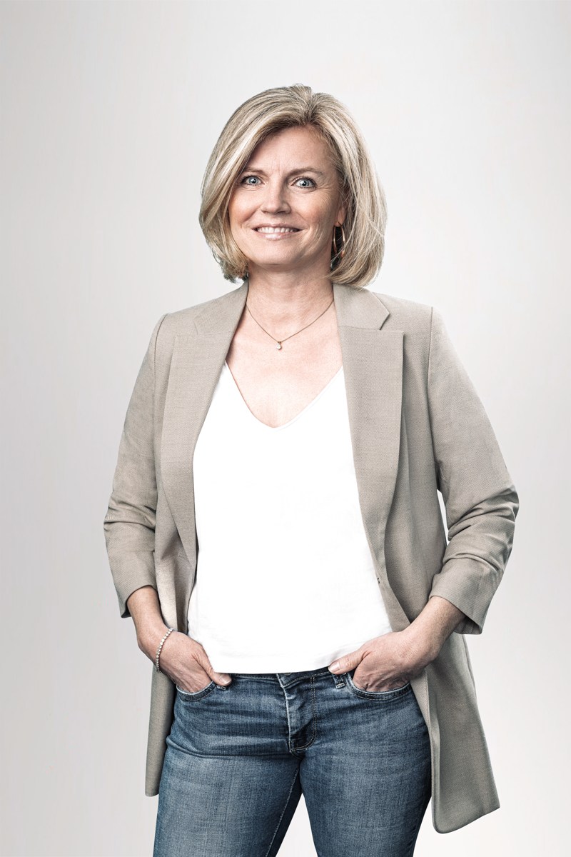 Eine lächelnde Frau mit blonden, schulterlangen Haaren trägt einen grauen Blazer über einem weißen Oberteil und Jeans. Sie steht mit den Händen in den Taschen und hat einen selbstbewussten Ausdruck. Der Hintergrund ist hell und neutral.