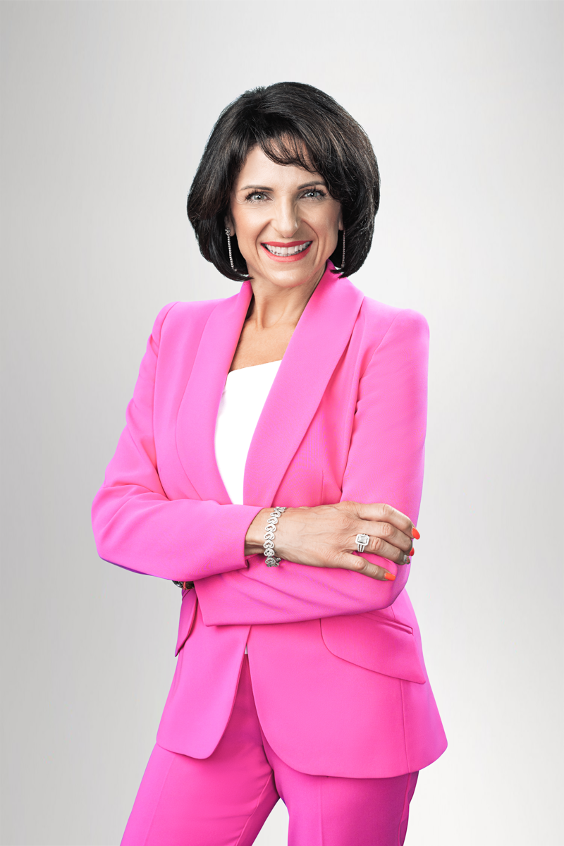 Eine Frau in einem auffälligen pinken Anzug, die mit verschränkten Armen und einem freundlichen Lächeln in die Kamera schaut. Der Hintergrund ist hell und neutral.