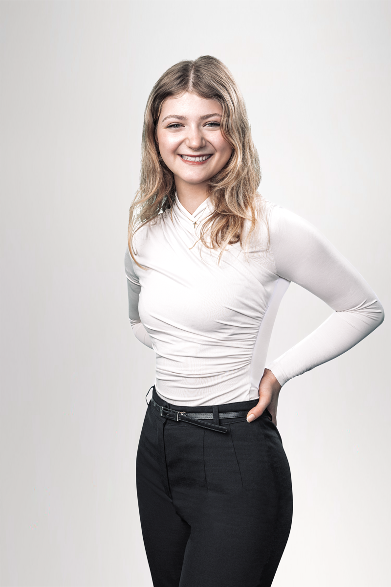 Eine lächelnde junge Frau mit langen, gewellten Haaren trägt ein eng anliegendes, weißes Oberteil und schwarze Hose. Sie steht mit einer Hand am Rücken und hat einen selbstbewussten Ausdruck.
