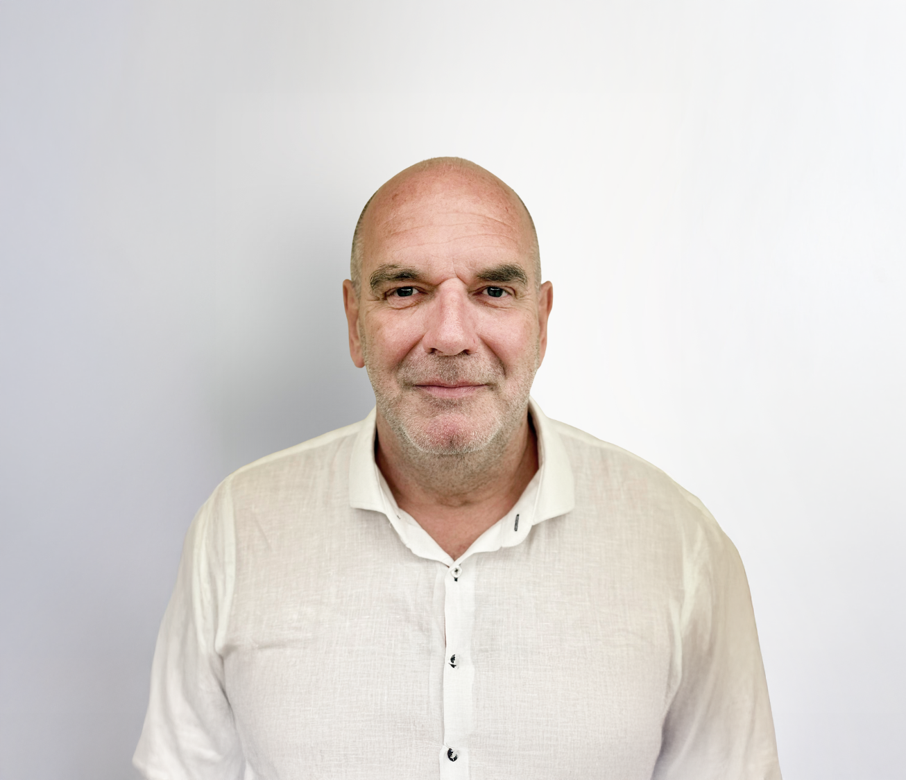 Ein mittelalter Mann mit Glatze und einem leichten Lächeln trägt ein helles, offenes Hemd. Der Hintergrund ist hell und neutral.