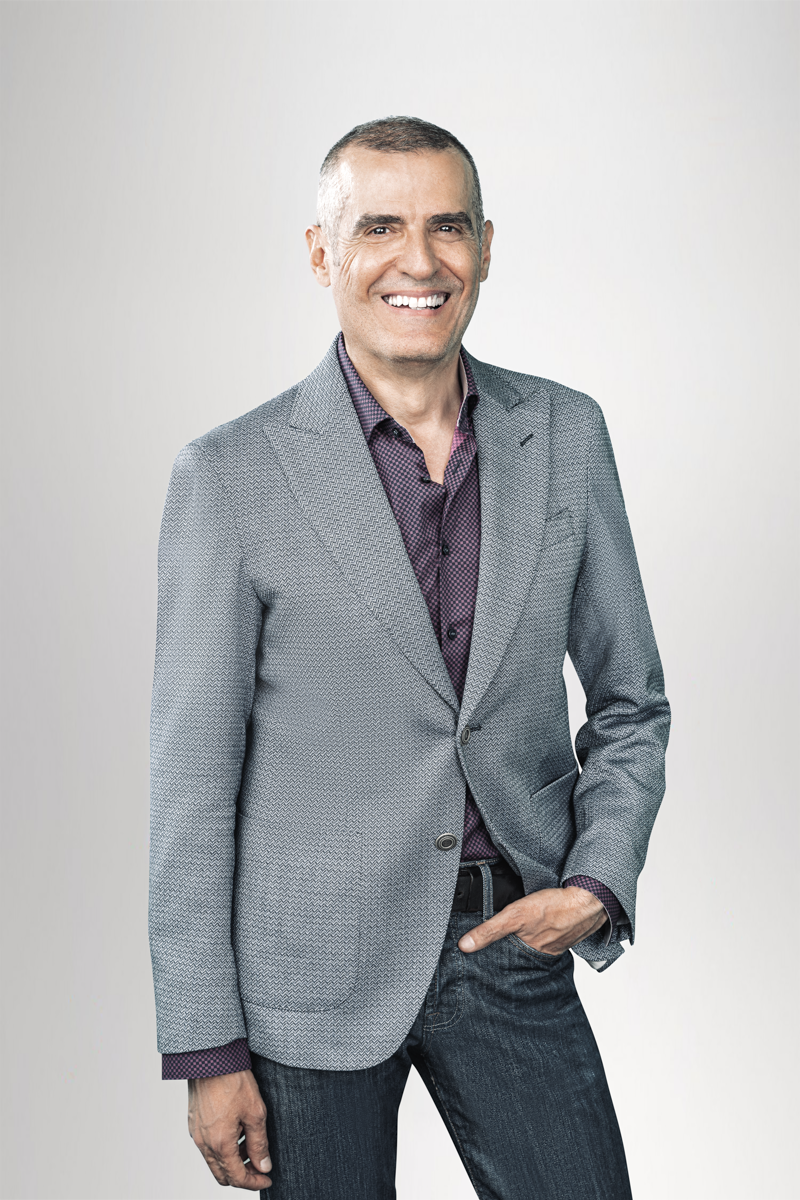 Ein lächelnder Mann mit kurzen, grauen Haaren trägt einen hellgrauen Blazer über einem lila Hemd und steht aufrecht vor einem neutralen, hellen Hintergrund.