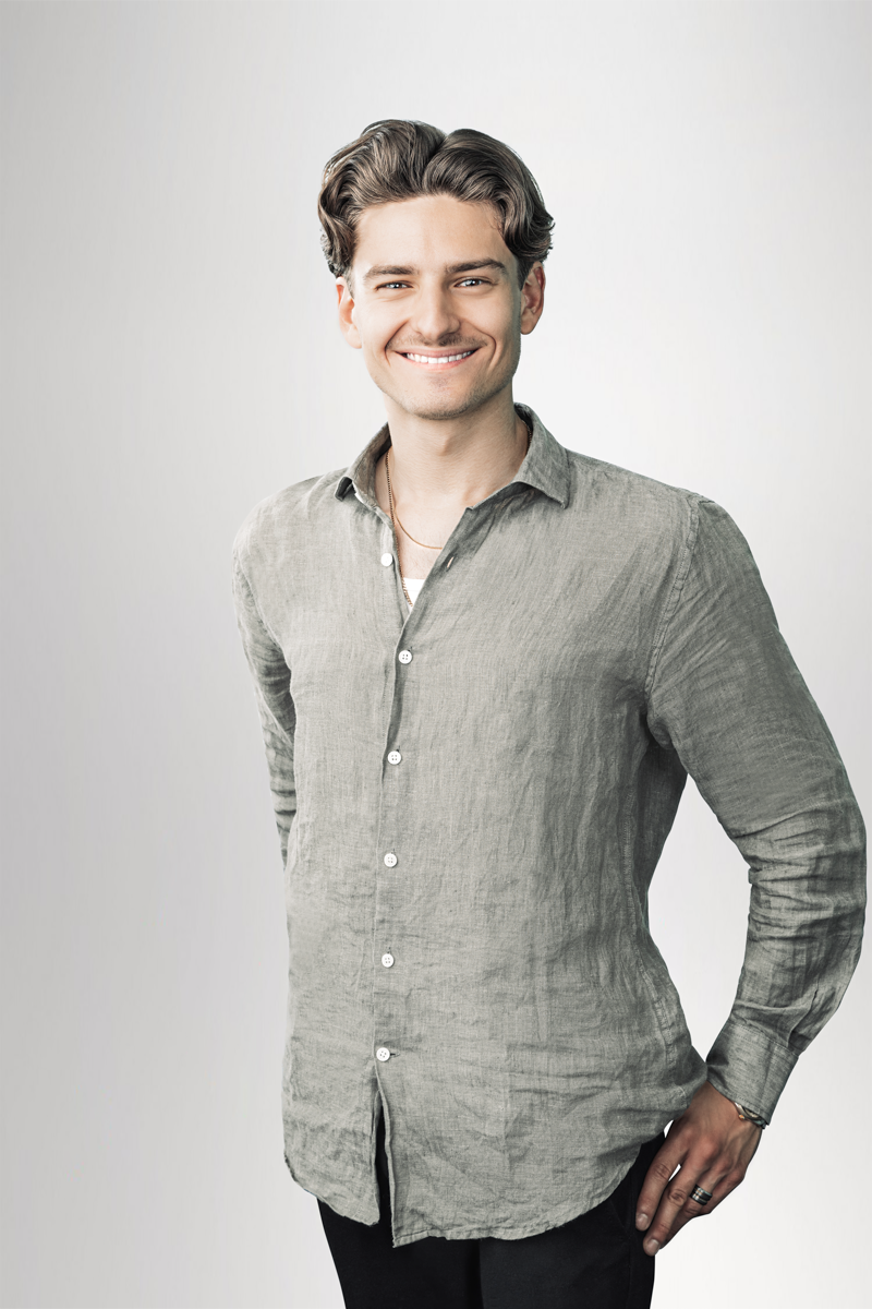 Ein junger Mann mit einem freundlichen Lächeln steht in einem grauen, leicht strukturierten Hemd vor einem hellen Hintergrund. Er hat lockige, schulterlange Haare und trägt mehrere Halsketten sowie Ringe. Seine Körperhaltung ist entspannt und selbstbewusst.