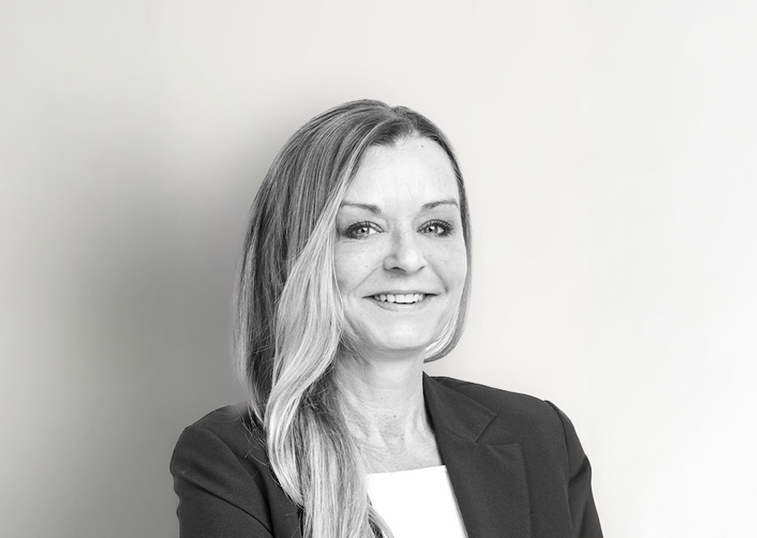 Eine Frau mit langen, glatten Haaren und einem freundlichen Lächeln trägt einen Blazer und steht vor einem hellen, einfarbigen Hintergrund.