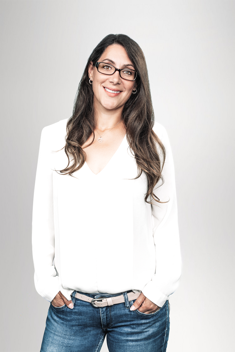 Eine lächelnde Frau mit langen braunen Haaren und Brille trägt ein weißes Oberteil und Jeans, während sie lässig mit beiden Händen in den Hosentaschen steht. Der Hintergrund ist hell und einfarbig.