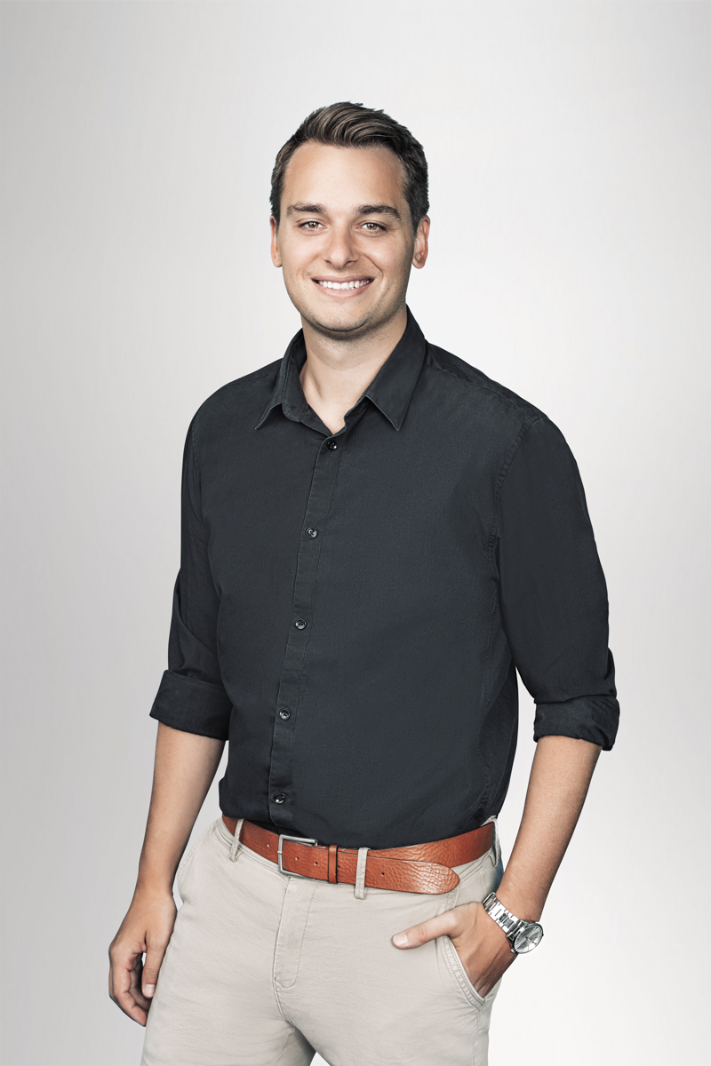 Ein junger Mann mit kurzes, dunkles Haar steht lächelnd vor einem neutralen Hintergrund. Er trägt ein schwarzes Hemd und eine helle Hose, ergänzt durch einen braunen Gürtel und eine Armbanduhr.