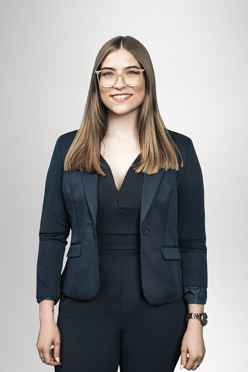 Eine Frau mit langen, glatten Haaren und Brille steht lächelnd vor einem hellen, neutralen Hintergrund. Sie trägt einen dunkelblauen Blazer über einem schwarzen Oberteil und hat eine Uhr an ihrem Handgelenk.