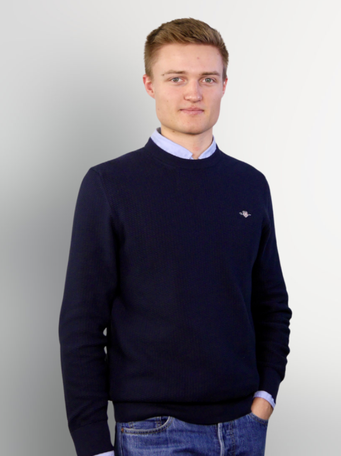 Ein junger Mann trägt einen dunkelblauen Pullover über einem hellblauen Hemd und steht mit verschränkten Händen vor einem hellen, einfarbigen Hintergrund.