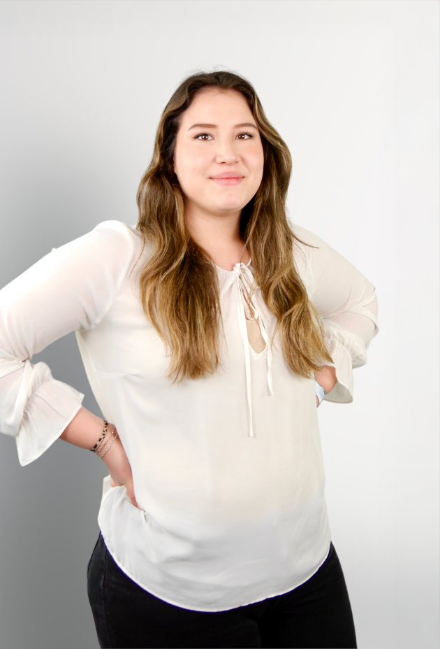 Eine junge Frau mit langen, gewellten Haaren steht lächelnd mit verschränkten Armen vor einem hellen, neutralen Hintergrund. Sie trägt eine blusige, cremefarbene Shirt mit einem Ausschnitt, der mit einer Schnürung verziert ist.