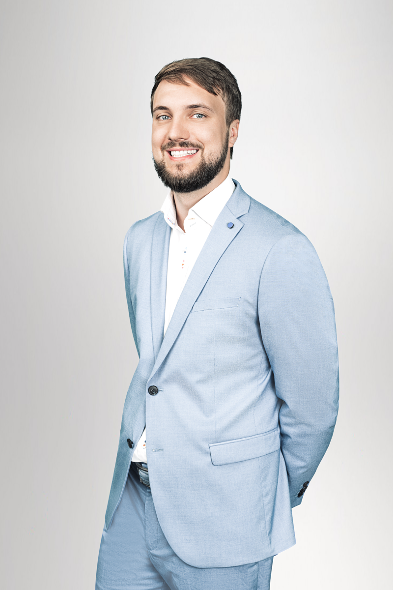 Ein lächelnder Mann in einem hellblauen Anzug steht mit einer Hand hinter seinem Rücken und blickt zur Kamera. Der Hintergrund ist hell und unifarben.