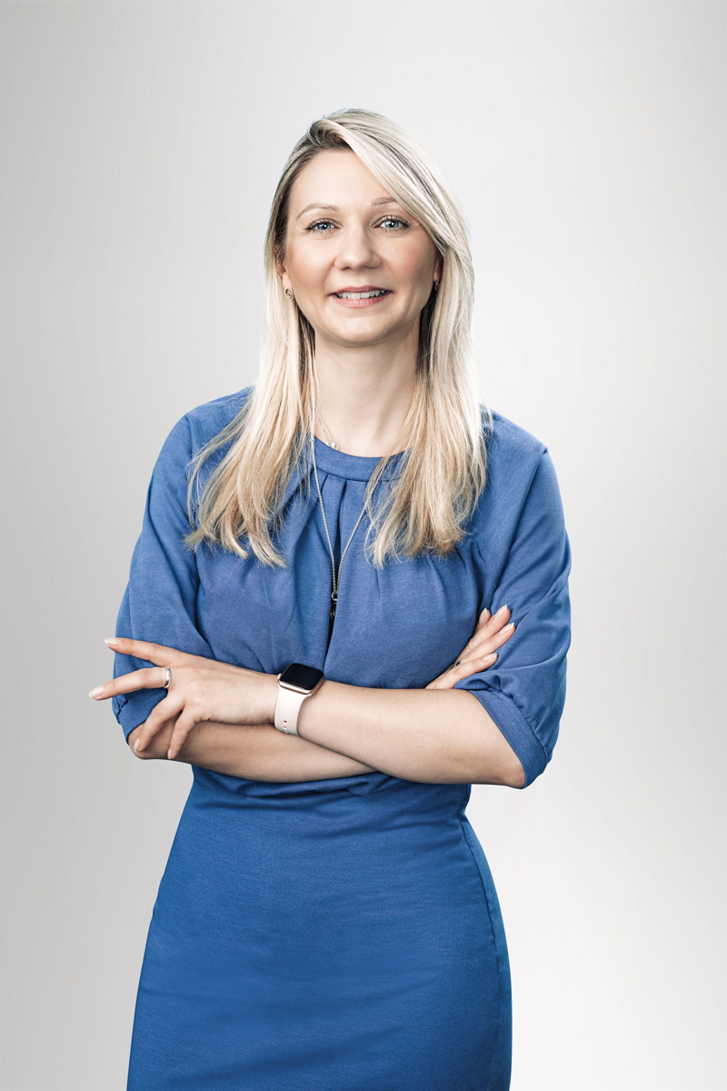 Eine lächelnde Frau mit langen, blonden Haaren trägt ein blaues Kleid und steht mit verschränkten Armen vor einem neutralen, hellen Hintergrund.