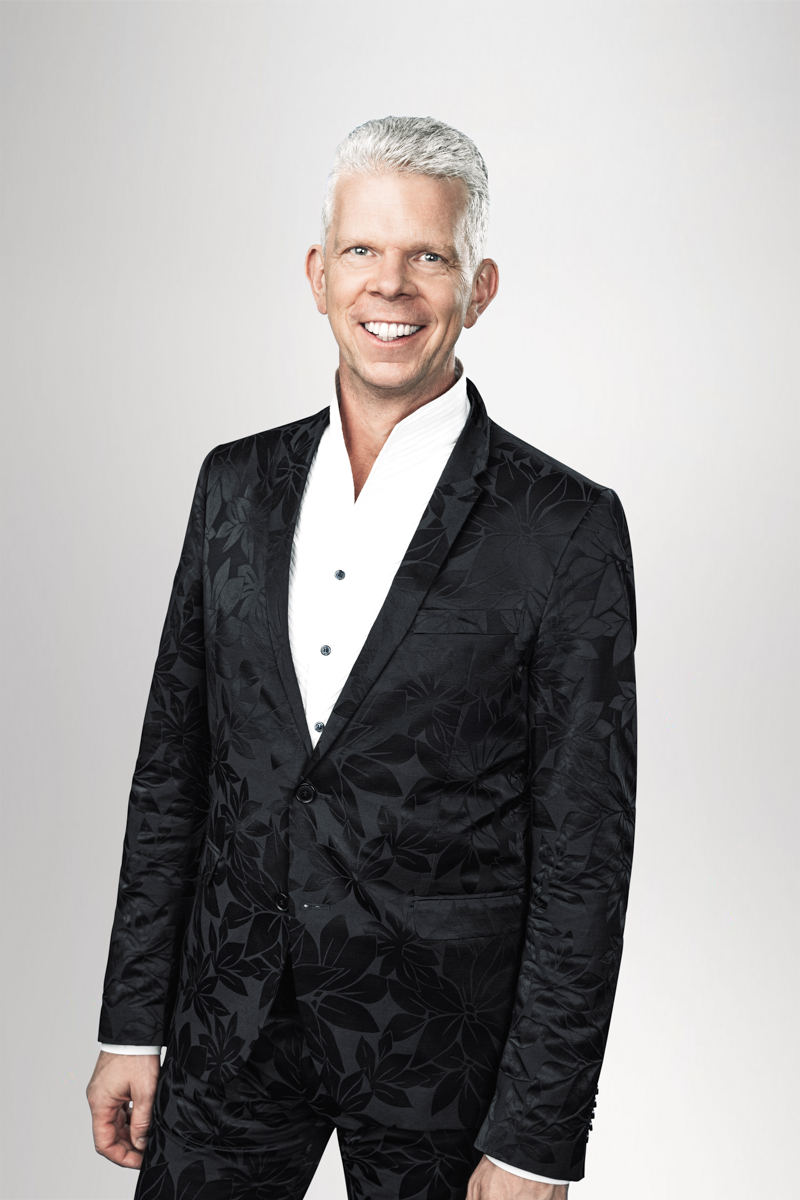 Ein lächelnder Mann in einem eleganten, schwarz gemusterten Anzug mit einem weißen Hemd. Der Hintergrund ist hell und neutral gehalten.